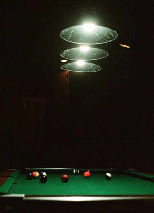 A green pool table with scattered balls under three hanging lamps in a dark room